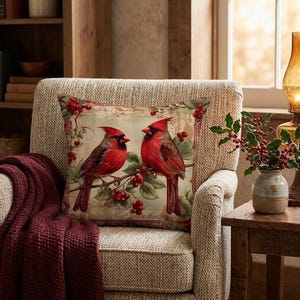 May include: A square throw pillow featuring a printed illustration of two red northern cardinal birds perched on a branch with green leaves and red berries against a light beige background with faint script text details.