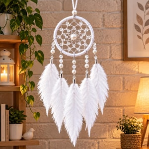 May include: A white dreamcatcher featuring a circular web woven with clear beads and a central pearl-like bead. Five strands hang from the hoop, each decorated with white beads and ending in a long, soft white feather. The item hangs by a cord.