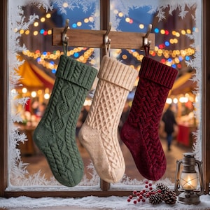 Può includere: Tre calze natalizie in maglia appese a un gancio di legno. Le calze sono verde bosco, crema e bordeaux scuro, ognuna caratterizzata da un motivo a trecce lavorato a maglia con un risvolto piegato nella parte superiore davanti a una finestra.