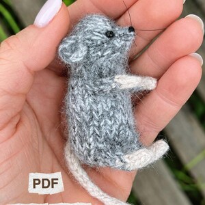 May include: A small, hand-knitted grey mouse figurine with a fuzzy texture, featuring cream-colored paws and a long tail, with black bead eyes and thin whiskers. The image includes white text labels that read PDF and KNITTING PATTERN.