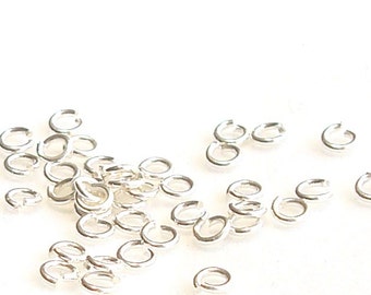 May include: A group of silver jump rings, scattered on a white surface. The rings are small and round, with a smooth finish.