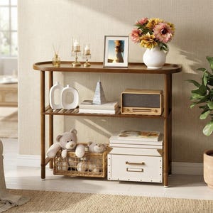 May include: A brown wooden console table with two shelves, featuring a top surface with a raised edge, holding a white vase with flowers, a picture frame, and candles. The lower shelves contain white ceramic decor, books, a radio, a basket with plush toys, and a white storage box.