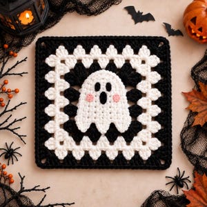 May include: A square crochet granny square featuring a central white ghost motif with black eyes and mouth and pink cheeks. The square is bordered by alternating rows of black and white yarn in a classic cluster stitch pattern on a flat surface.