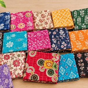 May include: A collection of folded square fabric pieces featuring various colourful patterns including floral, geometric, and circular motifs in shades of red, blue, yellow, green, purple, and black, all finished with visible white stitched quilting lines.