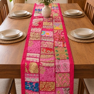Op de afbeelding: Een rechthoekige tafelloper met een patchworkontwerp van diverse roze, gouden en veelkleurige stoffen vierkanten. De loper is versierd met ingewikkeld borduurwerk, metaaldraad en kraalversieringen, afgewerkt met een effen roze rand.