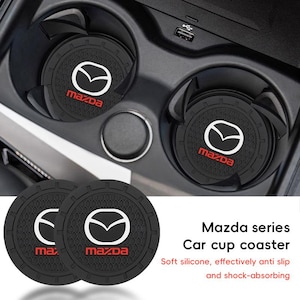 May include: Two circular black silicone car cup coasters featuring the white Mazda logo and red Mazda text. The coasters have a textured honeycomb pattern and a notched outer rim. The text Mazda series Car cup coaster is visible.