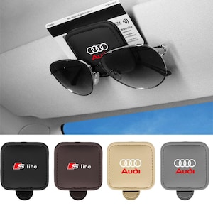 Könnte beinhalten: Ein schwarzer, quadratischer Sonnenblenden-Clip aus Leder mit weißem Audi-Logo, der eine dunkel getönte Sonnenbrille und eine Karte hält. Darunter befinden sich vier weitere Clips in Schwarz, Braun, Beige und Grau mit Audi- oder S-Line-Logo.