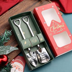 May include: A set of two silver-toned metal ice cream shovels with shovel-shaped heads and D-shaped handles, presented in a green gift box. Each shovel is engraved with the text 'Evie's Ice Cream Shovel'. The box features a red lid with 'Merry Christmas' text.