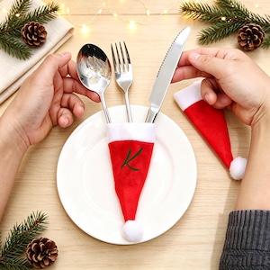 Può includere: Un set di posate in acciaio inossidabile composto da cucchiaio, forchetta e coltello seghettato inserito in un portaposate in feltro rosso a forma di cappello di Babbo Natale con bordo bianco, pon pon e lettera K ricamata in verde.
