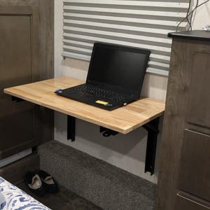 May include: A light-colored wooden wall-mounted folding desk with black metal support brackets. A black laptop is placed on the surface of the desk. The desk is positioned against a wall with horizontal blinds and is flanked by wooden cabinetry.