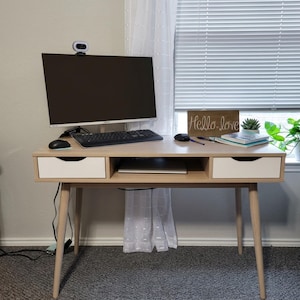 May include: A light wood desk with two white drawers, a central open shelf, and four tapered legs. On the desk sits a computer monitor with a webcam, a black keyboard, a black mouse, a wooden sign reading Hello love, a small succulent, and a notebook.