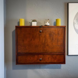 May include: A wall-mounted wooden drop-leaf desk featuring a rich brown finish, a single lower drawer with a round metal knob, and a fold-down front panel with a matching knob and a small metal latch mechanism at the top center of the unit.