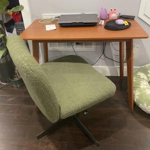 May include: A sage green textured fabric office chair with a swivel base and a wooden desk featuring a laptop, a black mouse pad, a purple mouse, and small decorative figures. The desk has four tapered wooden legs and a rectangular surface.