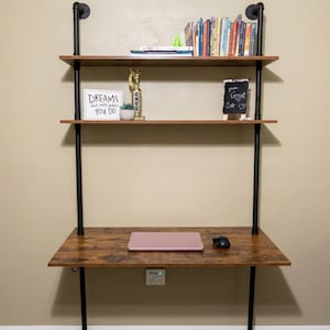 May include: A wall-mounted desk with two shelves featuring dark brown wood-grain surfaces and black metal pipe supports. The desk holds a pink laptop and a black mouse. Shelves contain books, a trophy, a succulent, and signs reading DREAMS and You're Great.