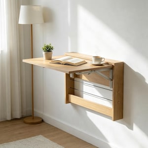 May include: A wall-mounted, light oak wood folding desk featuring a rectangular surface supported by a metal bracket mechanism. The desk is attached to a vertical panel with horizontal wire accents, holding an open book and a white ceramic coffee cup.