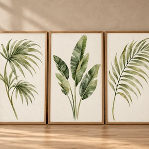 May include: A set of three rectangular wall art prints in light wood frames. Each print features a watercolor illustration of green palm and banana leaves on a cream-colored background, showcasing detailed botanical foliage.