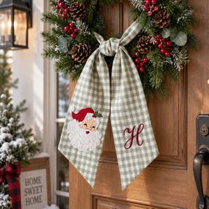 May include: A Christmas wreath featuring green pine needles, pinecones, and red berries, adorned with a sage green and white gingham ribbon. The ribbon features an embroidered Santa Claus face on one side and a red cursive letter H on the other.