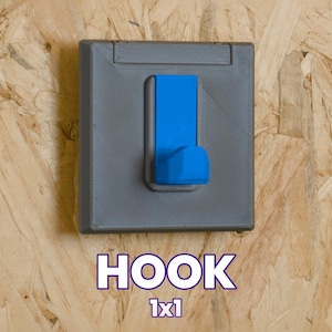 Puede incluir: Una base cuadrada gris montada en la pared con un gancho azul brillante en el centro. La base cuenta con una sección superior con bisagras. Debajo del objeto, el texto dice HOOK 1x1 en letras blancas con un borde oscuro, indicando el tipo y tamaño.