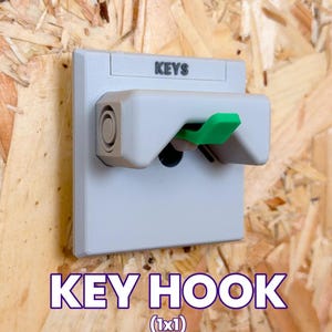 Puede incluir: Un gancho para llaves de pared de color gris claro con una base cuadrada y una carcasa rectangular que sobresale. La palabra KEYS está impresa en negro en la parte superior. Una palanca verde brillante sobresale del centro de la carcasa.
