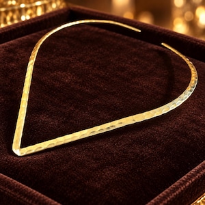 May include: A gold-toned V-shaped collar necklace featuring a textured, hammered surface. The necklace has an open design with tapered ends, creating a sharp, angular silhouette that rests in a point at the front, showcasing a metallic finish.