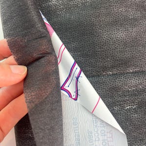 May include: A sheet of black non-woven fabric with a textured surface, partially peeled back to reveal a white backing layer. The white layer features printed blue and pink graphical outlines of a garment pattern with text in a mirrored orientation.