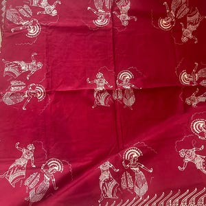 May include: A deep red fabric featuring a repeating white line-art pattern of figures in traditional dance poses. The textile includes a decorative border with a stylized leaf or flame motif along the top and bottom edges of the rectangular cloth.