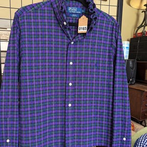 May include: A long-sleeved button-down shirt in a purple, green, and white plaid pattern. The shirt features a ruffled collar, a front button placket, and a Polo by Ralph Lauren label. A brown paper tag with the number 0165 is attached to the collar.