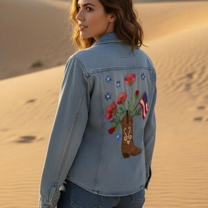 May include: A light blue denim jacket featuring an embroidered design on the back. The embroidery depicts a brown cowboy boot filled with red and pink flowers, accompanied by a small flag with red, white, and blue stripes and blue floral accents.