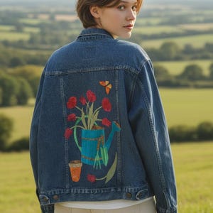 May include: A dark blue denim jacket featuring a painted design on the back. The artwork depicts a bright blue watering can filled with red tulips, accompanied by a small terracotta pot, a green trowel, and a small orange butterfly above.
