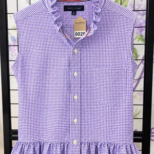 May include: A sleeveless Tommy Hilfiger button-down shirt in a purple and white gingham check pattern. The shirt features a ruffled collar, a ruffled hem, and a single chest pocket. A brown paper tag with the text 0026 is attached to the collar.