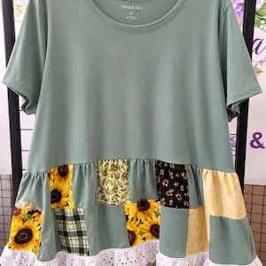 May include: A sage green short-sleeve t-shirt with a round neckline and a label reading Terra Sky 1X. The lower section features a patchwork design of sunflowers, green leaves, black floral, and plaid patterns, finished with a white eyelet lace trim.