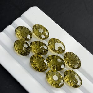May include: A collection of nine oval-shaped, faceted olive-green gemstones arranged in a grid pattern on a white, ridged display tray. Each stone features a distinct radial cut pattern that reflects light, creating a shimmering, translucent appearance.
