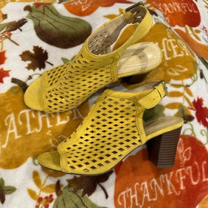 May include: A pair of yellow suede slingback sandals featuring a laser-cut diamond pattern across the vamp, an open toe, and a stacked block heel. Each sandal includes an adjustable ankle strap with a metal buckle and the Liz Claiborne brand name.