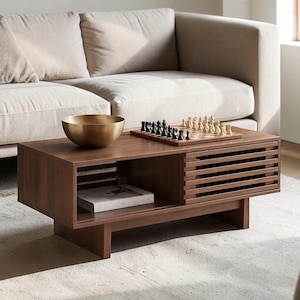 May include: A rectangular dark wood coffee table with a slatted sliding door, an open storage shelf containing a book titled GATHERING, a brass bowl, and a wooden chess set with black and white pieces arranged on the tabletop surface.