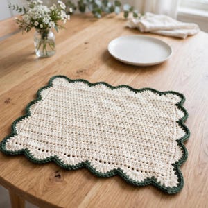 May include: A rectangular crochet placemat featuring a cream-coloured centre with a textured stitch pattern and a scalloped border in a dark forest green yarn. The placemat has a flat, woven construction with a decorative, wavy edge finish.