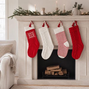 May include: Four Christmas stockings hanging from a mantel. The first is red with a white snowflake pattern and cuff. The second is solid white with a cable knit texture. The third has red and white horizontal stripes. The fourth is solid red cable knit.