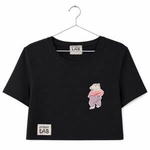 Op de afbeelding: Een zwart kort T-shirt met korte mouwen, een ronde halslijn en een metalen hanger. Het shirt toont een illustratie van een capibara in een roze shirt en paarse broek, met een OFFBEAT LAB label bij de nek en aan de rechteronderkant.