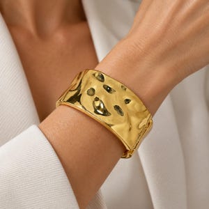 May include: A wide, gold-toned cuff bracelet featuring a highly reflective, polished surface with an irregular, hammered texture. The bracelet has a substantial width and a hinged design, showcasing a smooth, metallic finish that catches the light.
