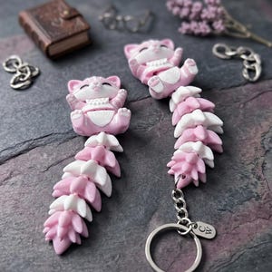 May include: Two articulated cat figurines made of pink and white material. Each cat has a head with closed eyes and a long, segmented tail consisting of alternating pink and white pieces. One cat is attached to a silver metal keyring with a small tag.