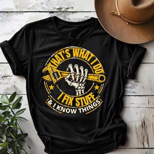 May include: A black short-sleeved t-shirt featuring a graphic of a skeleton hand holding a yellow adjustable wrench. The design includes the text "THAT'S WHAT I DO I FIX STUFF & I KNOW THINGS" in yellow lettering arranged in a circular pattern.