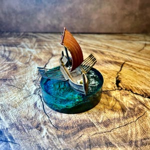 May include: A miniature model of a wooden ship with a curved brown sail and multiple oars on each side, mounted on a circular base of translucent blue resin that mimics the appearance of deep ocean water with textured waves and foam.