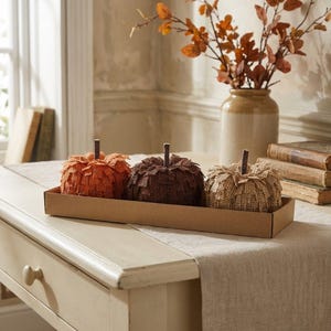 May include: Three decorative pumpkins crafted from textured hessian fabric in shades of orange, dark brown, and tan. Each pumpkin features a small, dark brown stem and is arranged in a single row inside a rectangular, light brown cardboard tray.