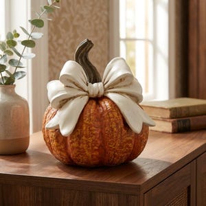 May include: A decorative orange pumpkin figurine with a textured, bumpy surface and a dark brown stem. A large, smooth, off-white bow is attached to the top of the pumpkin, resting just below the stem, creating a contrast between the colours.
