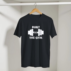 Pode incluir: Uma t-shirt preta de manga curta com um gráfico branco de uma barra de pesos centrado no peito. O texto BUILT FOR THE GYM está impresso em letras maiúsculas brancas, com a palavra FOR posicionada dentro do gráfico da barra.
