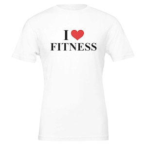Pode incluir: Uma t-shirt branca de manga curta com o texto I seguido de um símbolo de coração vermelho e a palavra FITNESS impressa em letras maiúsculas pretas no centro do peito. A peça tem gola redonda e uma bainha reta padrão.