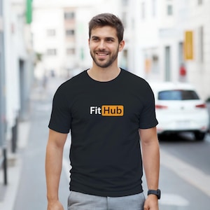Può includere: Una t-shirt nera a maniche corte con girocollo e la scritta Fit Hub stampata sul petto. La parola Fit è in bianco e la parola Hub è in bianco all'interno di un riquadro rettangolare arancione. La maglietta ha un taglio standard e un design semplice.
