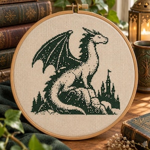 Può includere: Un telaio da ricamo circolare in legno contenente un disegno a punto croce su tessuto beige. Il motivo raffigura un drago verde scuro di profilo accanto alla sagoma di un piccolo castello, con una griglia testurizzata visibile.