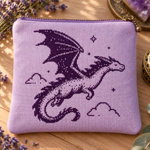 May include: A square lavender fabric pouch featuring a dark purple cross-stitch embroidery of a flying dragon. The design includes small star and cloud motifs surrounding the dragon. The pouch has a metal zipper closure at the top edge.