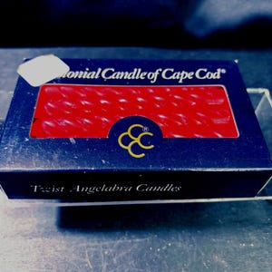 May include: A rectangular dark blue box labeled Colonial Candle of Cape Cod containing red twist Angelabra candles. The box features a central cutout window displaying the candles, a gold logo with three interlocking Cs, and white text.