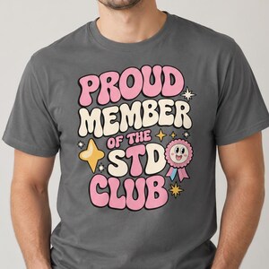 May include: A charcoal gray short-sleeve t-shirt featuring a graphic print with the text PROUD MEMBER OF THE STD CLUB in pink and white bubble letters. The design includes yellow four-pointed stars and a pink ribbon award with a smiling face.